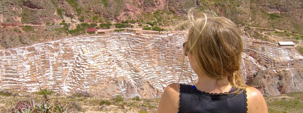 The view of the salt mines from across the valley is just as spectacular. Best Hikes In Peru Image: A woman with a blonde braid pulled over her right shoulder is wearing a black top, sunglasses, and is observing the salt mines from across the valley.