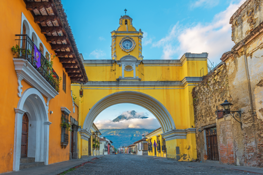Belize and Guatemala Itinerary Image: Yellow Spanish-Colonial arch on a street in Antigua, with mountains seen through the archway.