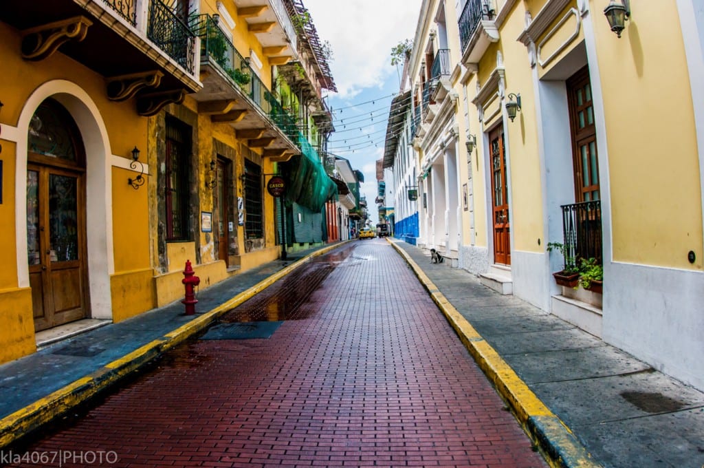 Colourful buildings always seem to have an added charm. Architecture In Panama Image: A narrow city street is lined with buildings on both the left and the right. We can see shades of mustard yellow, pale lemon, turquoise, and marine blue gracing their faces. Many have wrought iron balconies.
