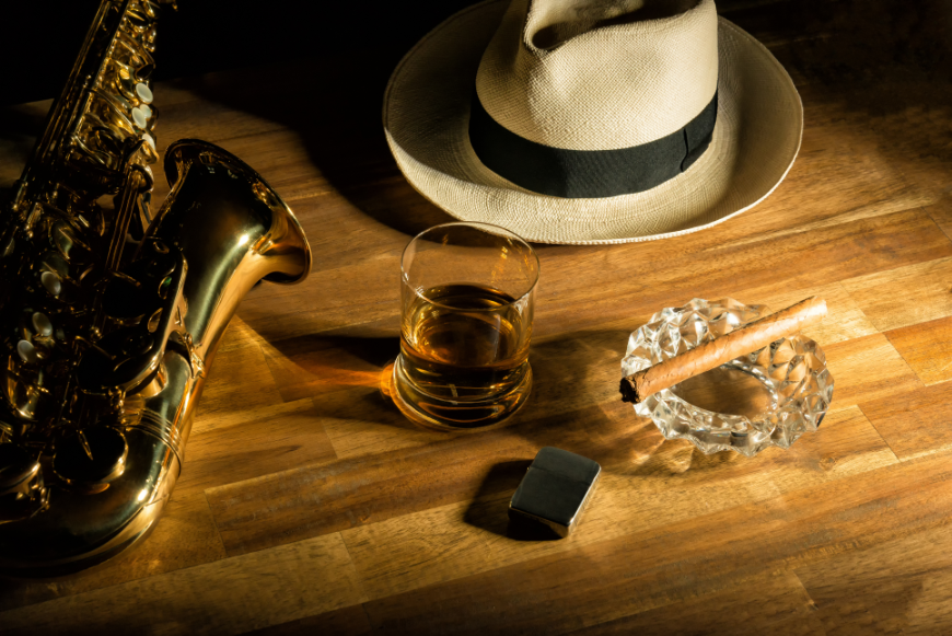Salsa Dancing in Cuba Image: A Panama hat, saxophone, glass of rum, silver lighter, and cigar perched on crystal ashtray sit on a wooden bar.